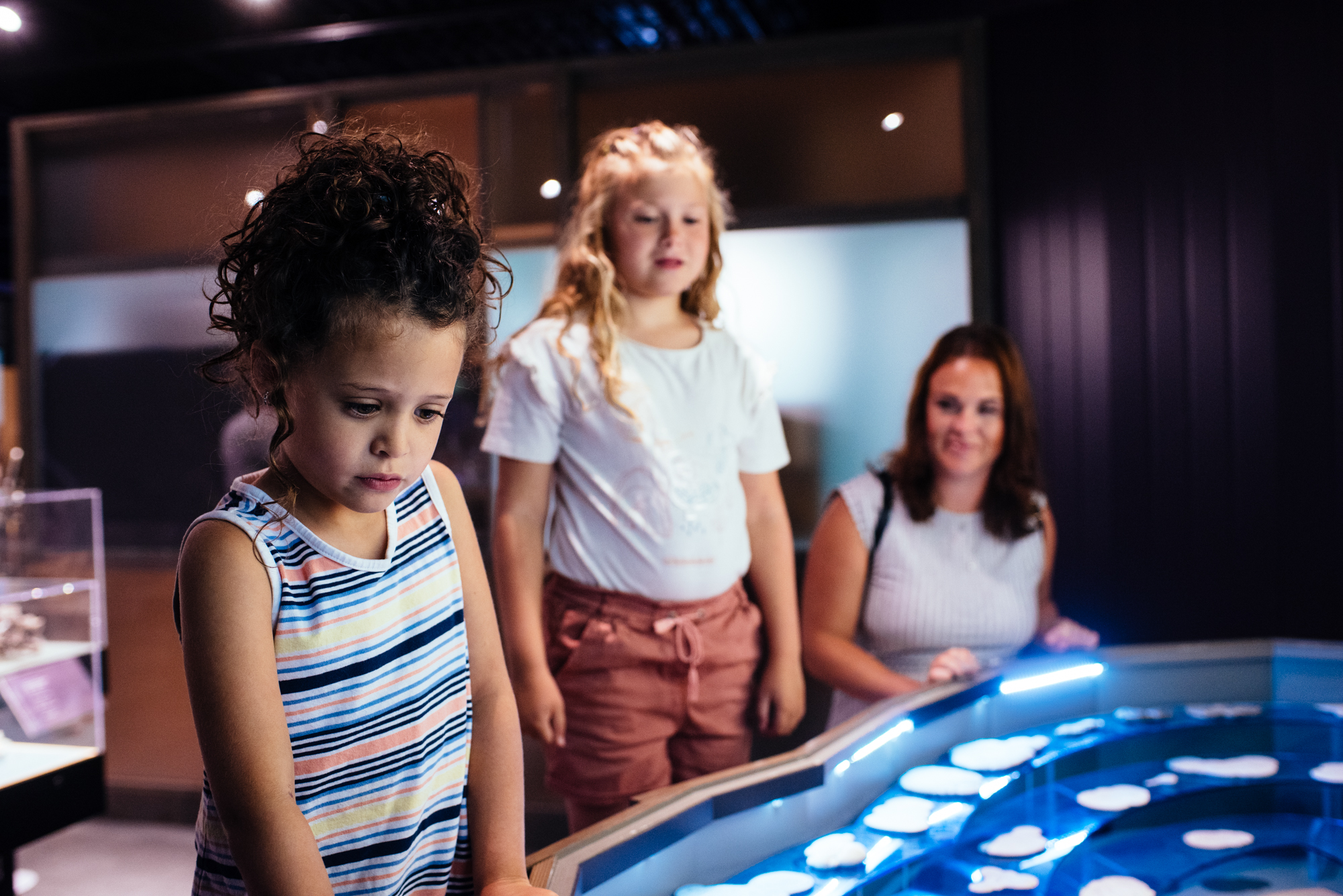 Deux enfants regardent un kiosque de pierres de fée au Musée minéralogique de Malartic sous le regard bienveillant de leur maman. Visiter le musée minéralogique est une belle activité familiale à faire en Abitibi-Témiscamingue. 